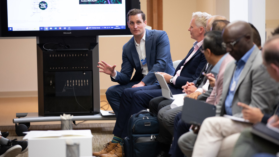 A group of people seated in a semi-circle in an office or conference room, engaged in a discussion. One person is speaking and gesturing with their hand while others listen. A large screen in the background displays presentation content.